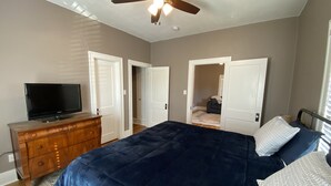 Quarto com cama grande, cômoda, televisão e ventilador de teto. Duas portas dão acesso a outros espaços.