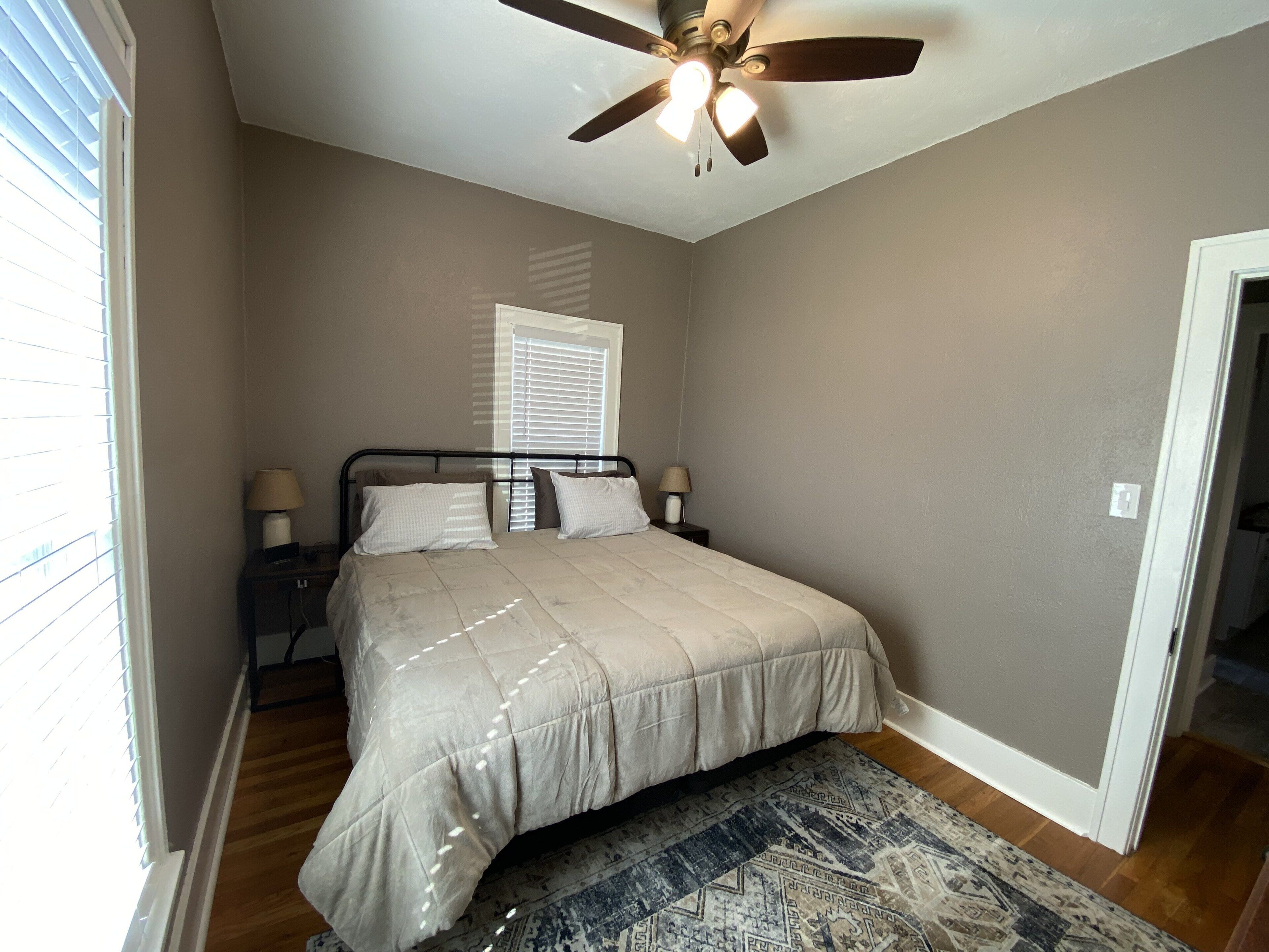 A bedroom with a bed, two bedside tables with lamps, a ceiling fan, a window with blinds, and a patterned rug.