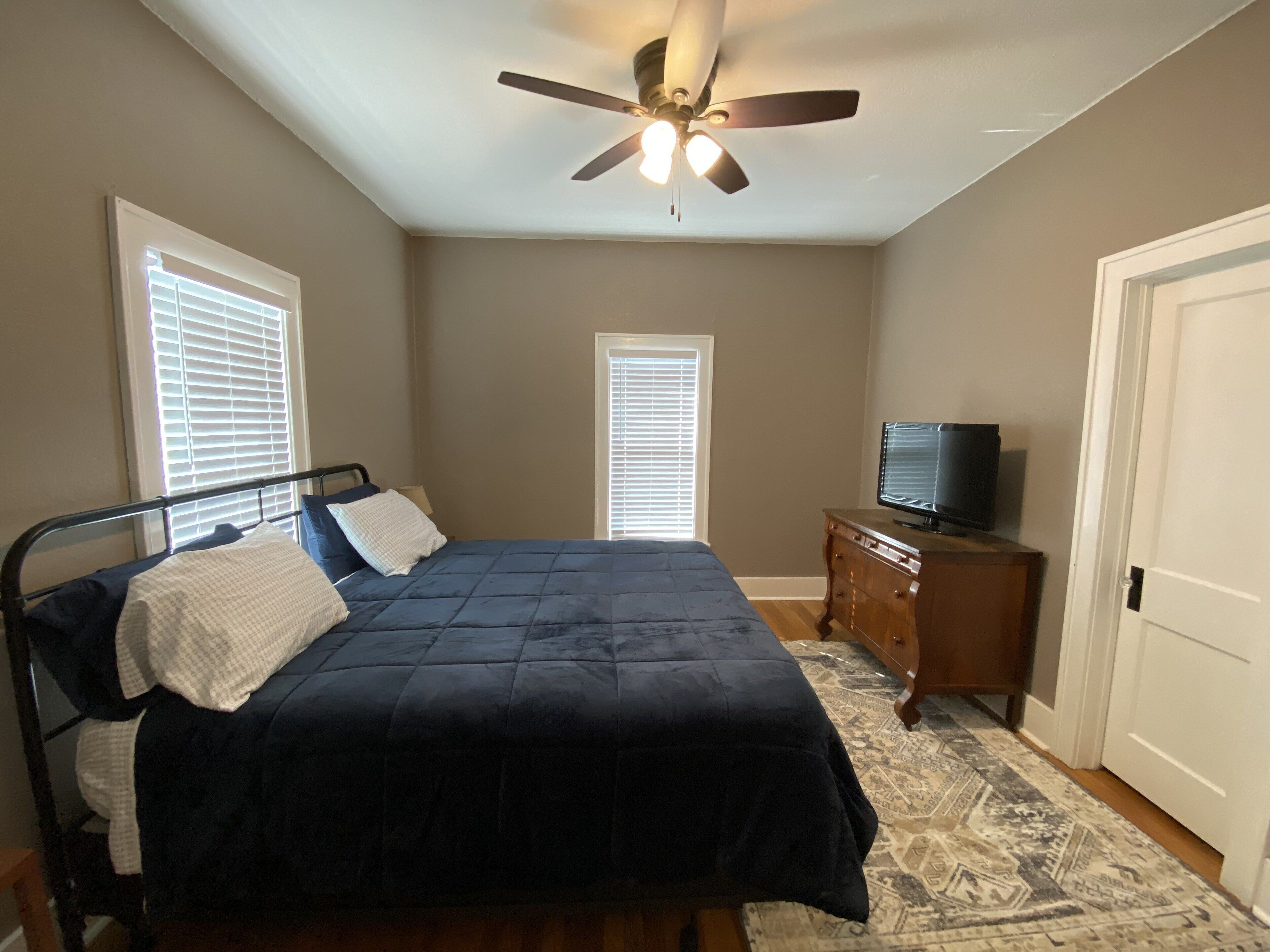 Une chambre à coucher avec un lit, un ventilateur de plafond, un téléviseur posé sur une commode et un tapis à motifs.