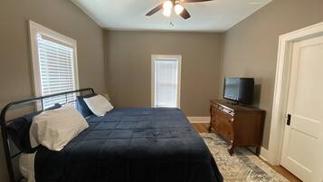 Une chambre à coucher avec un lit, un ventilateur de plafond, un téléviseur posé sur une commode et un tapis à motifs.