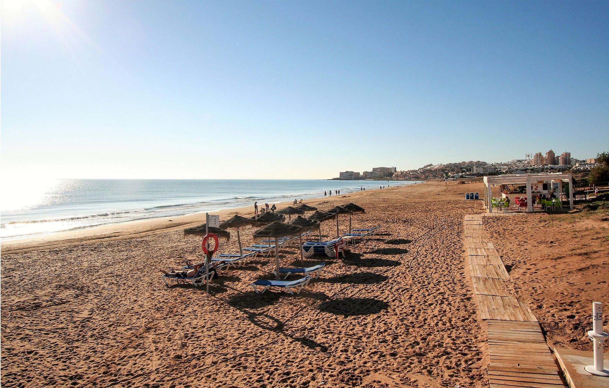 A beach with sun loungers and umbrellas, a wooden walkway, and a distant cityscape.