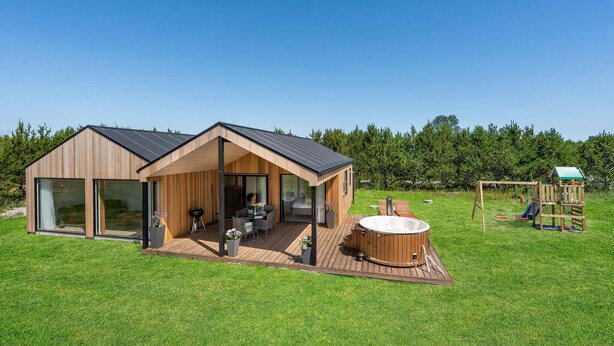 Eine moderne Holzblockhütte mit großer Veranda, Whirlpool und Kinderspielplatz auf einer Rasenfläche.