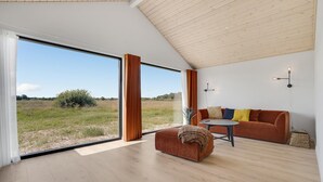 Ein geräumiger Raum mit einer großen Schiebeglaswand, die zu einem Feld im Freien führt, einer Holzbalkendecke und einer gemütlichen Sitzecke mit Sofa und Hocker.