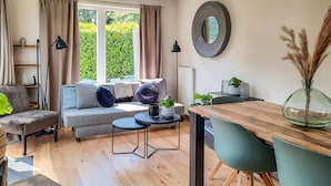 Een moderne woonkamer met een bank, een eettafel en een salontafel. Er is een raam met gordijnen, een lamp en een spiegel aan de muur.