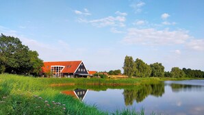 Een traditioneel Nederlands huis met een rood dak, gelegen naast een rustig meer met weelderige begroeiing en bomen op de achtergrond.