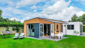Een modern huis met een grote glazen deur, een terras met buitenmeubilair en een houten hek op de achtergrond.