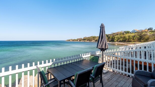 A deck with a table and chairs overlooking a clear blue sea, a white fence, and a house in the background. - Beach front home in Hasle (Hasle)