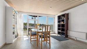 A dining area with a round table and chairs, a view through large windows, and a bookshelf with various items. - Beach front home in Hasle (Hasle)