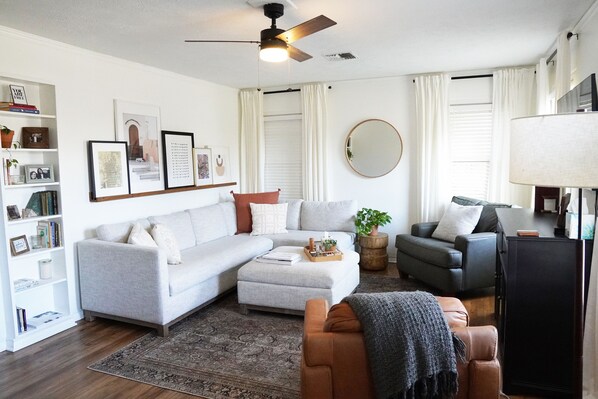 A living room with a sofa, armchair, and ottoman, a coffee table, a bookshelf, a ceiling fan, and a TV. - The Village House - 3 bedroom, 1 bath home 1.5 mi to Texas A&M, 2 mi to Legends (Bryan)