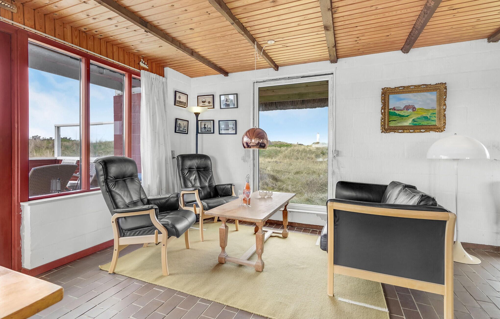 Une pièce chaleureuse avec un plafond en bois, de grandes fenêtres et une vue sur le paysage. On y trouve deux fauteuils en cuir noir, une table en bois et un canapé en cuir noir.