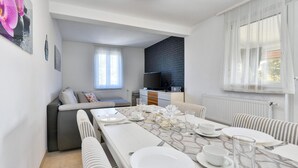 Un espace repas moderne avec une longue table dressée pour quatre personnes, un canapé, un téléviseur et une fenêtre ornée de rideaux légers.