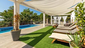 Ein Poolbereich mit Kunstgrasanlage, Liegestühlen und einer Pergola mit weißen Vorhängen.