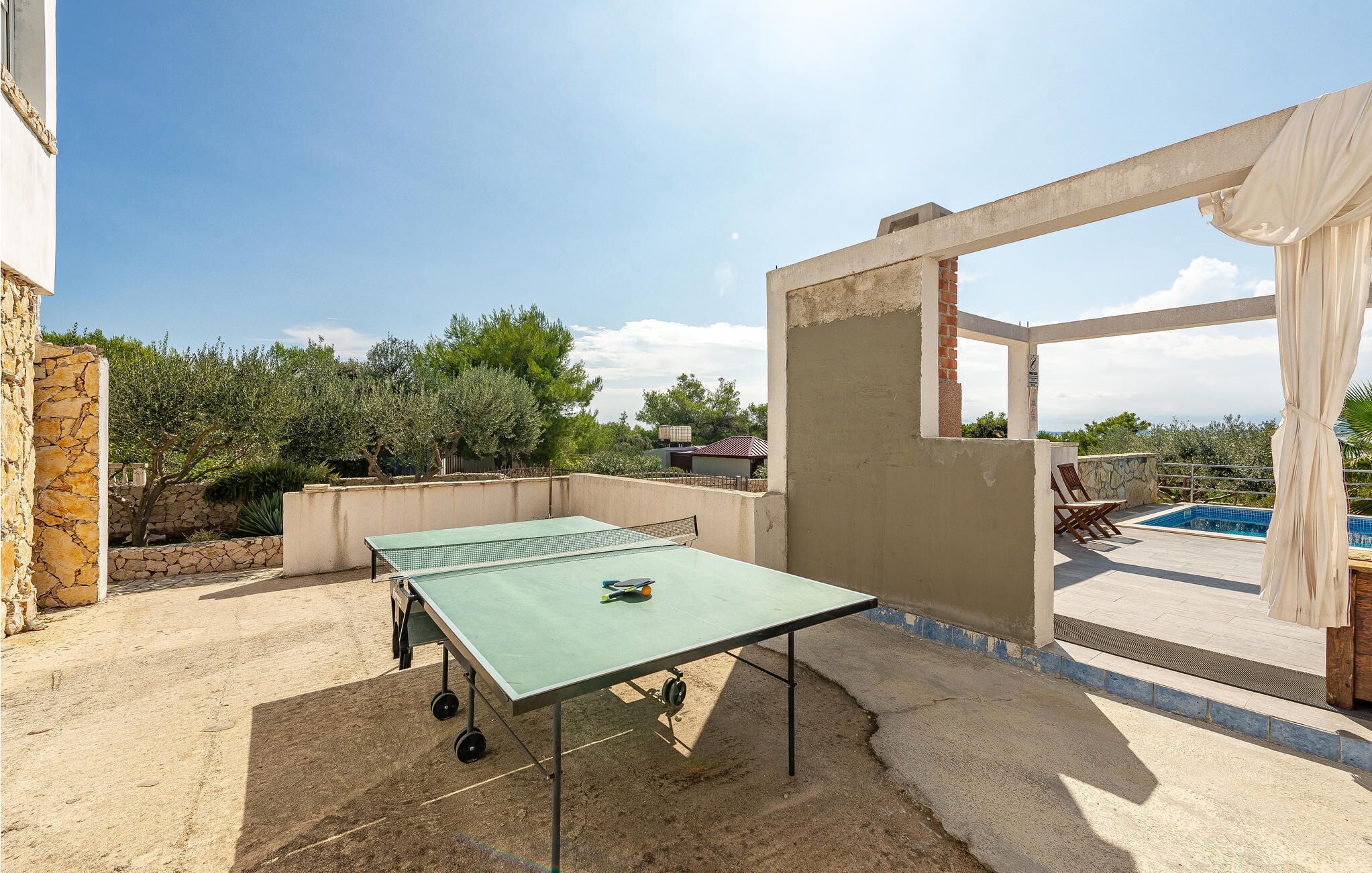 Mesa de tênis de mesa ao ar livre em um pátio com piscina e vista para árvores e edifícios ao fundo.