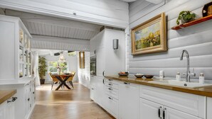 Une cuisine avec des meubles blancs, un plan de travail en bois, un tableau au-dessus de l’évier et un coin repas avec une table et des chaises.