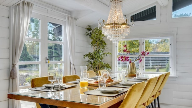 Un espace repas avec une table en bois, des chaises jaunes, un lustre et une vue sur la verdure à l’extérieur.