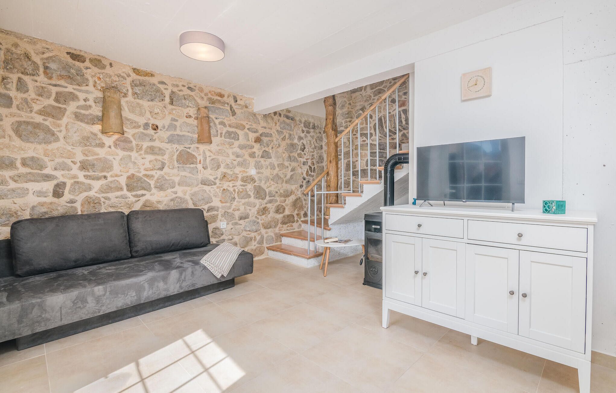 Un salon avec un mur en pierre, un canapé gris, un meuble blanc et un escalier.