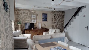 A cozy living room with a stone wall, a wooden TV stand, a sofa, a dining table with chairs, and a staircase. - House by the sea (Plouguerneau)