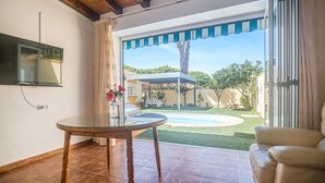 A room with a glass table, a sofa, a TV mounted on the wall, and a view of a pool and garden outside.