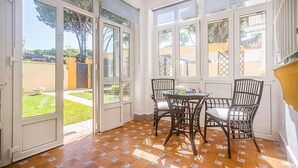 A sunlit room with a glass door leading to a garden, a table with chairs, and a tiled floor.