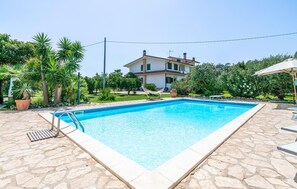 A rectangular swimming pool with a stone deck, surrounded by lush greenery and a house in the background.