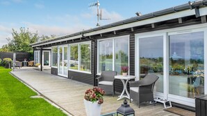 Ett modernt hus med stora glasdörrar, en träaltan, utemöbler och en liten vattendekoration.