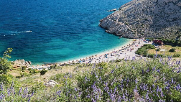 Una spiaggia costiera con acque cristalline turchesi, un litorale roccioso e un sentiero che conduce a una zona spiaggia con ombrelloni e lettini.