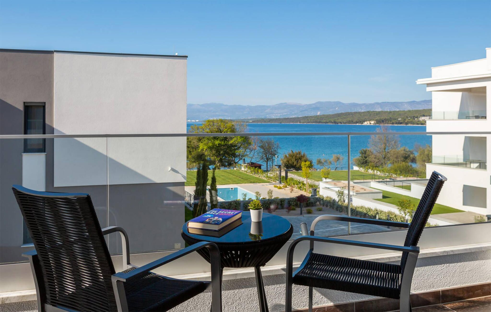 A balcony with two black chairs and a small table, overlooking a body of water and a landscaped area with trees and a pool.