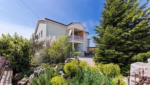 Un bâtiment à deux étages doté d’un balcon, entouré d’un jardin où poussent différentes espèces de plantes et bordé d’un mur en pierre.