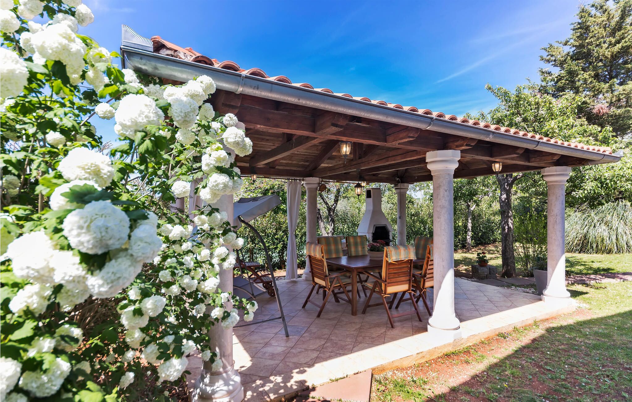A covered patio with a table and chairs, surrounded by white flowers and trees.