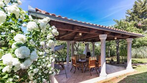 Une terrasse couverte avec une table et des chaises, entourée de fleurs blanches et d’arbres.