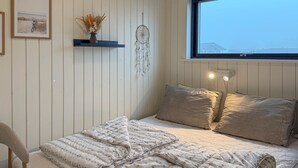 Un dormitorio con cama, almohadas, una lámpara de noche, un atrapasueños, un cuadro enmarcado y una ventana con vistas.
