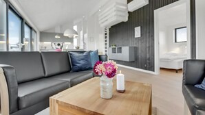 Una sala de estar moderna con un sofá gris, una mesa de centro de madera con un jarrón de flores y una vela, y una cocina al fondo.