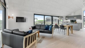 Una sala de estar moderna con sofá, mesa de comedor y sillas, una zona de cocina con armarios grises y ventanas amplias que ofrecen vistas al exterior.