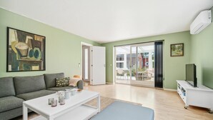 Ein Wohnzimmer mit einem grauen Sofa, einem weißen Couchtisch und einem blauen Hocker. Ein großes Schiebeglastür führt in einen Außenbereich mit Tisch und Stühlen.