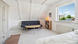 Ein Schlafzimmer im Loft-Stil mit schrägverdachter Decke, einem dunkelgrauen Sofa, einem kleinen runden Tisch, einem Bücherregal, einem Fenster mit Ausblick und einem Bett mit weißer Bettwäsche.