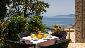 Una mesa lista para el desayuno con vista al mar, sillas de mimbre y una pared de piedra.