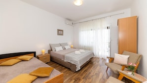 Un dormitorio con una cama grande, dos camas más pequeñas, un armario de madera, una silla con un jarrón de flores y una ventana con cortinas traslúcidas.