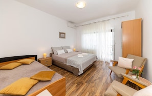 A bedroom with a large bed, two smaller beds, a wooden wardrobe, a chair with a vase of flowers, and a window with sheer curtains.