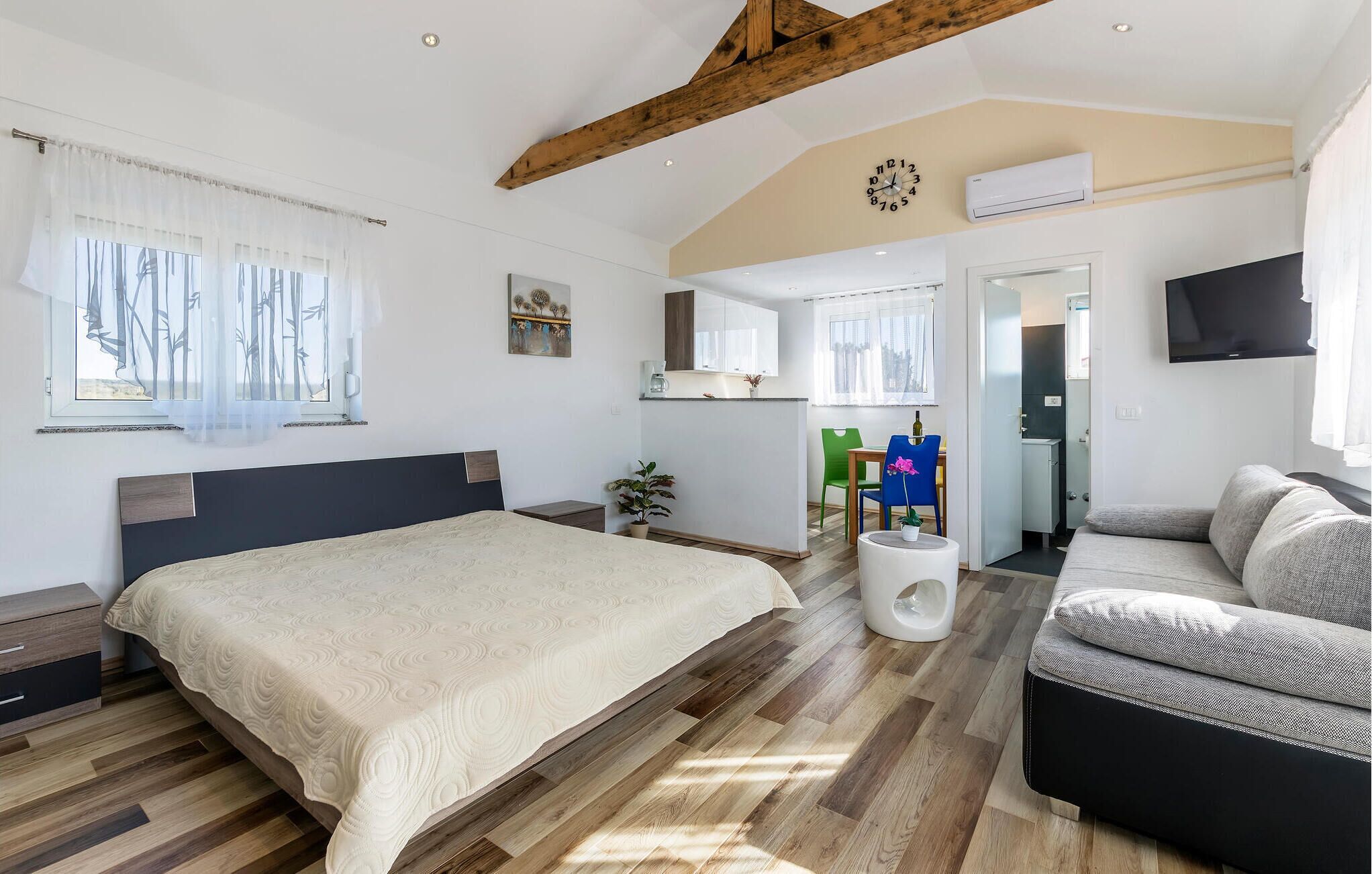 A spacious bedroom with a large bed, a sofa, a small table, and a kitchenette in the background.