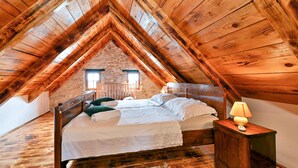 Ein gemütliches Schlafzimmer unter dem Dach mit Holzbalkendecke, einem Bett mit weißen Leinentüchern, einem Nachttisch mit Lampe und einer Steinwand.