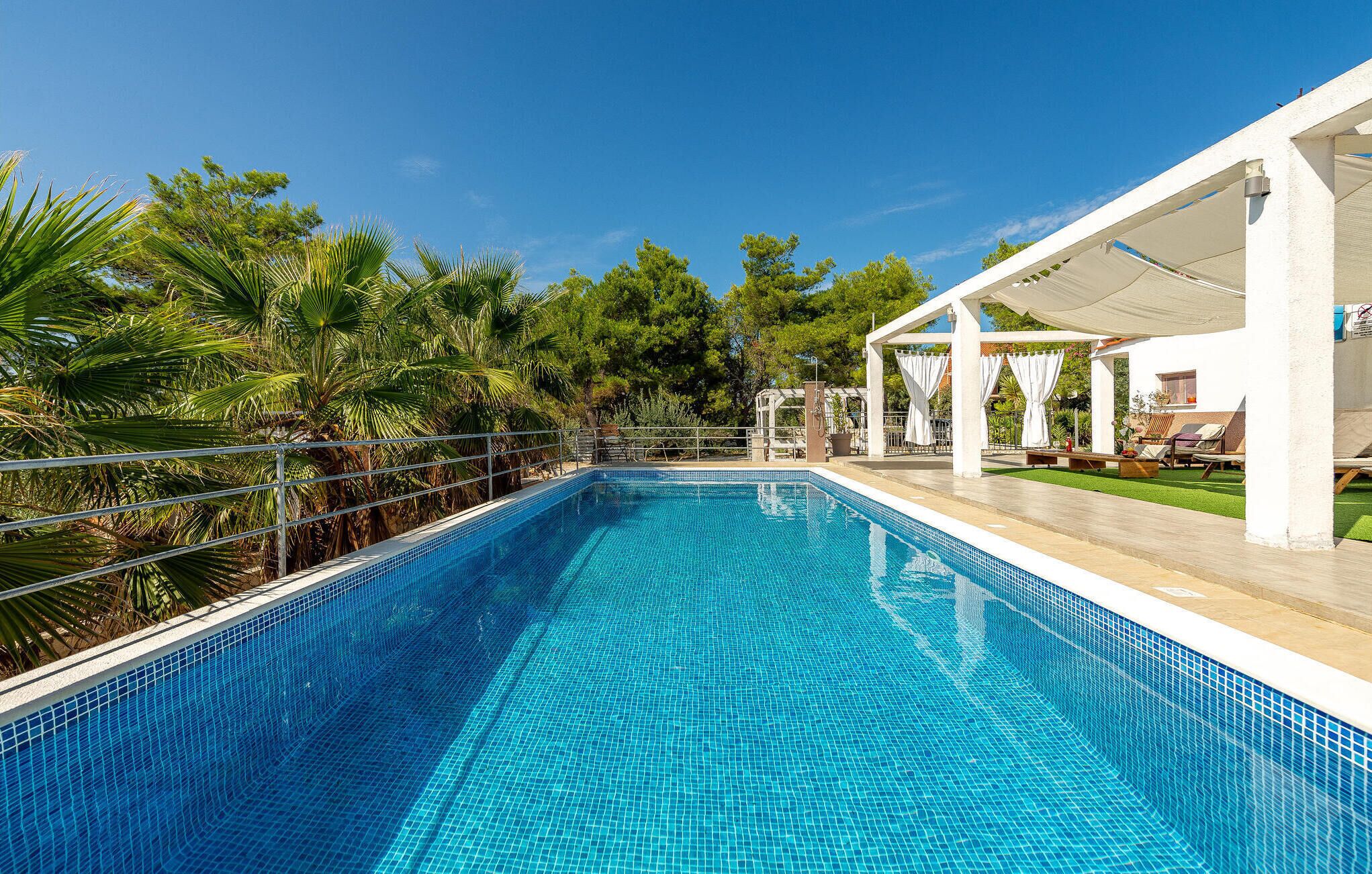 Ein rechteckiger Swimmingpool mit klarem, blauem Wasser, umgeben von einem Metallgeländer. Auf der rechten Seite befindet sich ein überdachter Sitzbereich mit weißen Markisen und Liegestühlen.