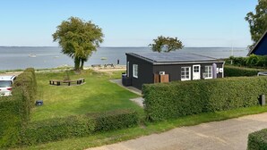 A small, dark-colored building with a solar panel roof, a grassy lawn, a tree, and a view of the sea. - Beach front home in Sjølund (Sjølund)