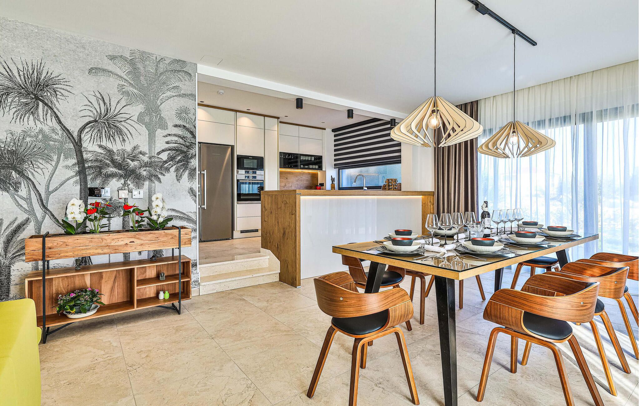 Una cocina moderna con una isla central, una zona de comedor con sillas de madera y un mural de palmeras en la pared.