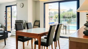 Un comedor moderno con una mesa y sillas de vidrio, un reloj en la pared y vista a una piscina a través de grandes puertas correderas.