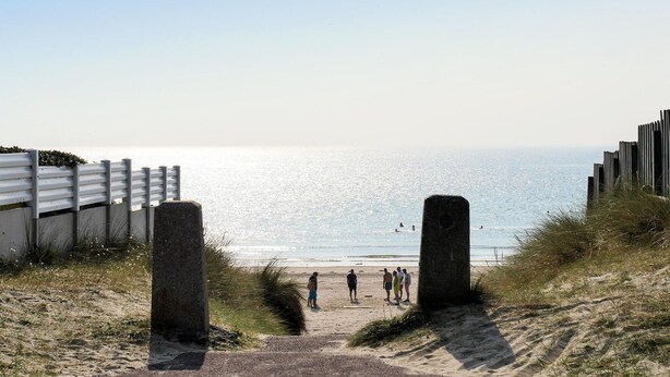 Una entrada a la playa con un sendero arenoso que conduce al mar, bordeada por estructuras de madera y un grupo de personas.