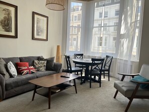 Una sala de estar con sofá, mesa de centro y mesa de comedor con sillas. Hay dos cuadros enmarcados en la pared, una lámpara y una ventana con cortinas.