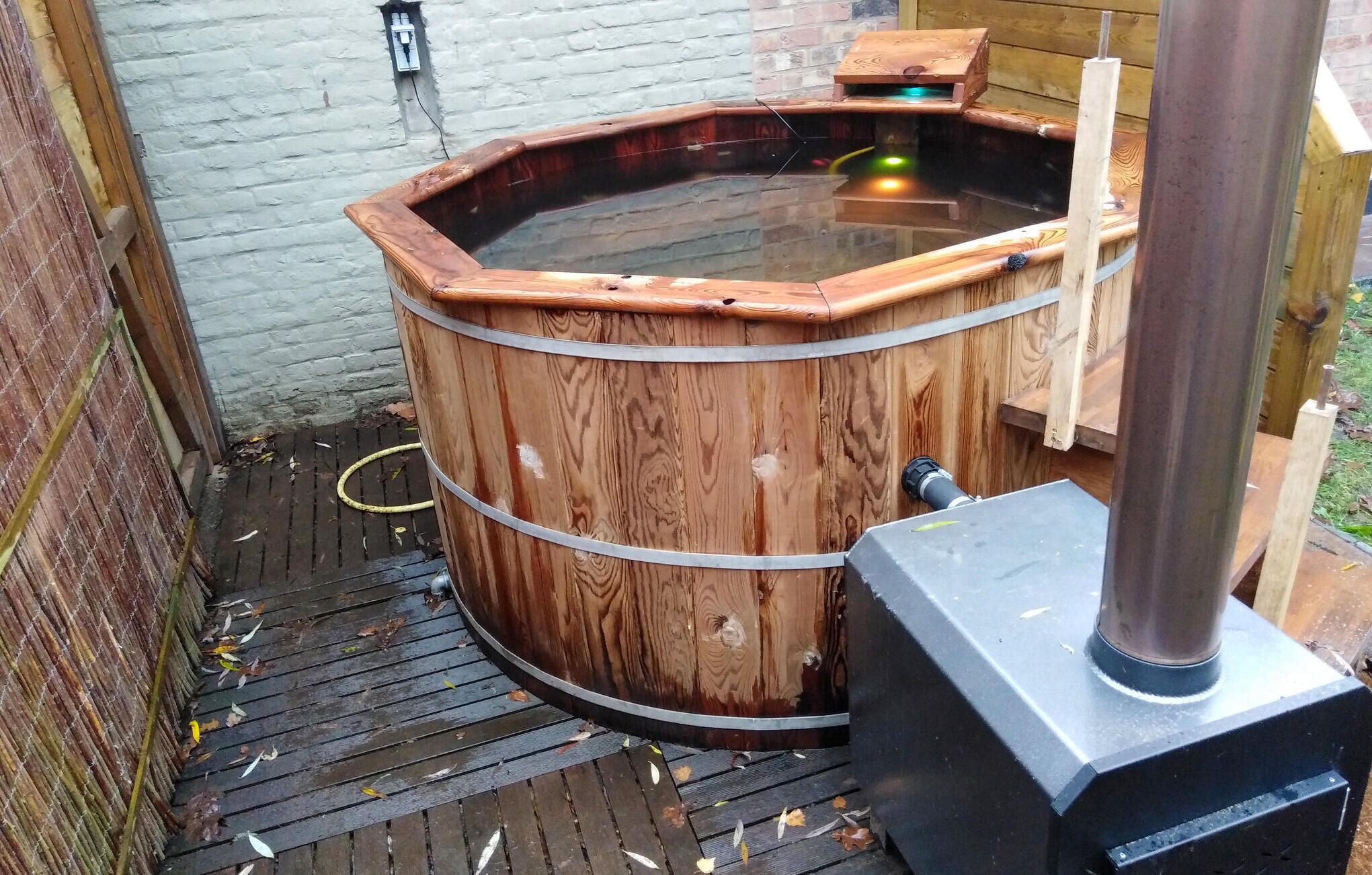 A wooden hot tub with a metal frame, a wooden deck, and a brick wall in the background.