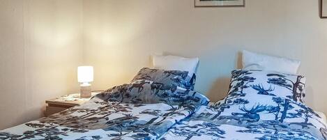 Een slaapkamer met een bed, een nachtkastje, een lamp en twee ingekaderde foto’s aan de muur.