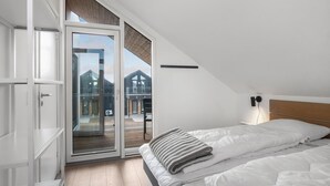 Un dormitorio con un amplio ventanal corredera que da a un balcón, una cama con ropa blanca y un cabecero de madera.