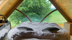Ein Schlafzimmer mit großem Fenster, einem Bett mit weißer Bettwäsche und Blick auf eine grüne Landschaft.